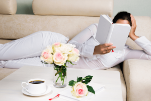 5 Life-Changing Benefits of Reading -The image depicts a woman dressed in white, reclining on a beige couch while reading a book. She is holding the book up, partially covering her face.