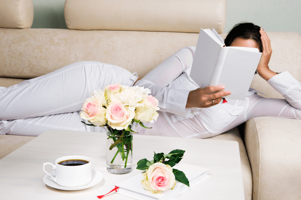 5 Life-Changing Benefits of Reading -The image depicts a woman dressed in white, reclining on a beige couch while reading a book. She is holding the book up, partially covering her face.